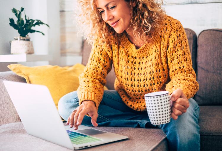woman sits on couch holding coffee cup looking at laptop